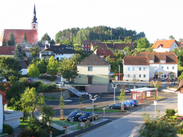 Das Bild zeigt den Dorfplatz Dechantskirchen mit Bushaltestelle, Parkplätzen und der Kirche im Hintergrund