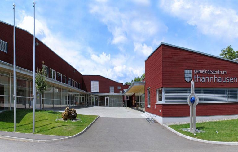 Gemeindezentrum Thannhausen. Ein Modernes Gebäude in Dunkelrot mit gepflastertem Platz im Zentrum