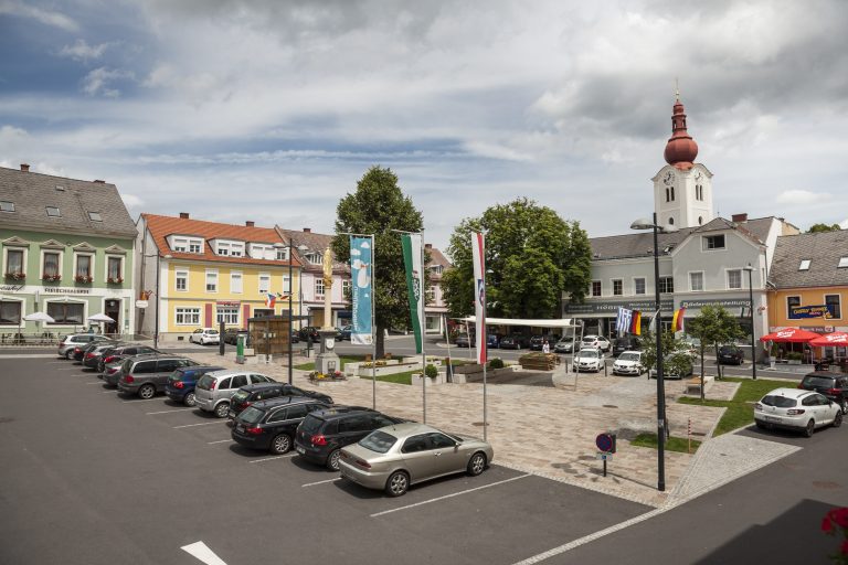 Das Bild zeigt den Friedberger Hauptplatz. In der Mitte des Platzes ist eine gepflasterte Fläche mit Sitzgelegenheiten und einem Sonnensegel. Rund um diese Fläche befinden sich zahlreiche Parkplätze