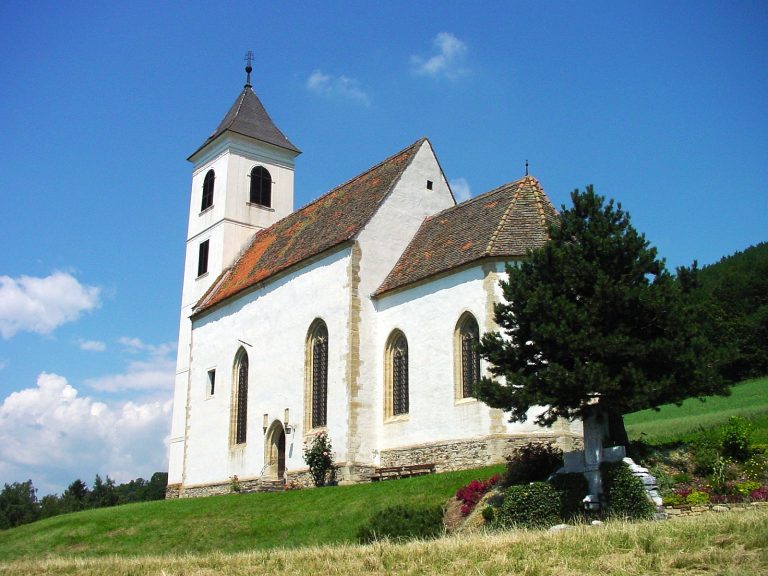 Das Bild zeigt die Kirche St. Anna. Die ist weiß und relativ schmal und steht an einem Hang umgeben von Wiesen