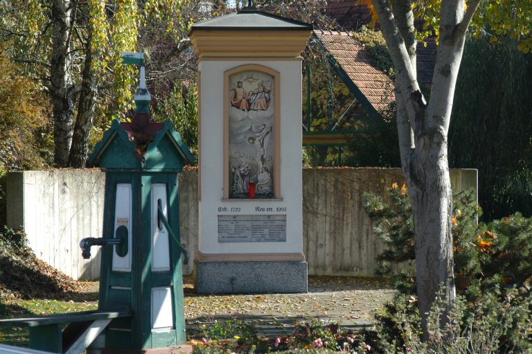 Dorfplatz in Pirching mit Bäumen, einem Brunnen sowie einem Denkmal