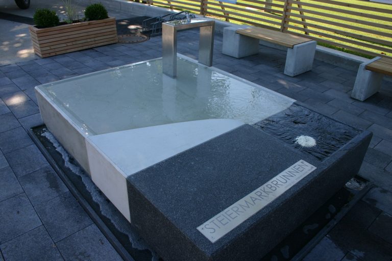 Das Bild zeigt einen modernen steinernen Brunnen mit der Aufschrift "Steiermarkbrunnen"