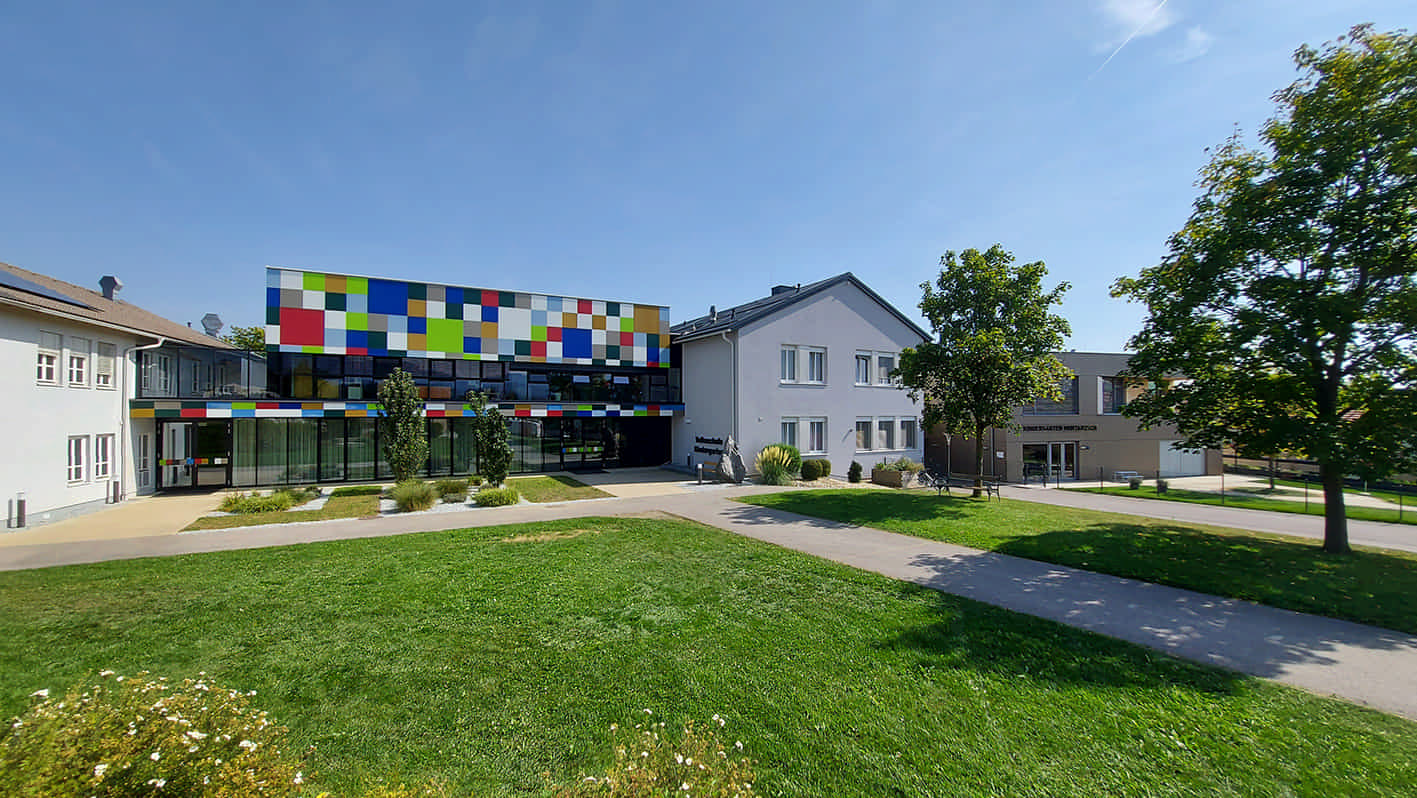 Gemeindezentrum Mortantsch mit kleinem Park im Vorfeld. Das Gebäudeensemble besteht aus Teilen unterschiedlicher Bauepochen. Im Zentrum steht ein Neubau mit Glasfront und bunten Kacheln als Fassade