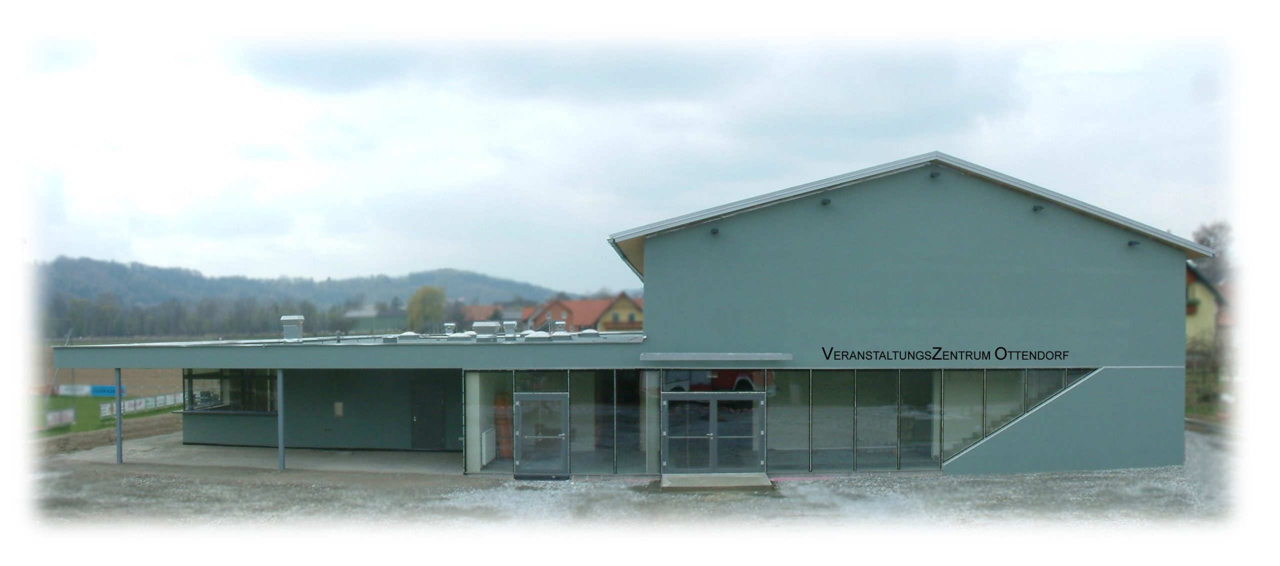 Veranstaltungszentrum Ottendorf. Graues Gebäude mit Glasfront am Ortsrand neben einem Sportplatz