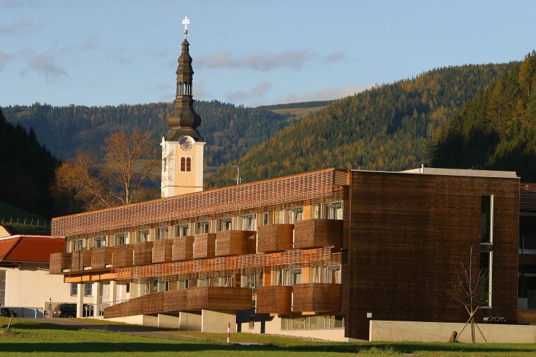 Das Bild zeigt das Pflegeheim Kamillus in Passail. Das zweistöckige Gebäude ist mit Holz verkleidet. Im Hintergrund ist der Kirchturm zu sehen