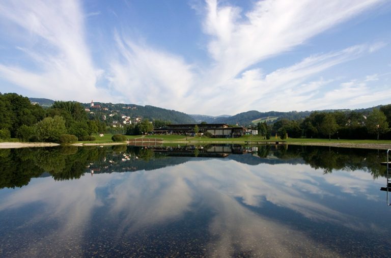 Im Pinggauer Badesee spiegeln sich die Wolken. Am gegenüberliegenden Ende befindet sich ein Restaurant