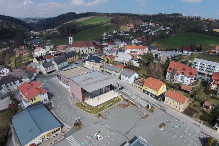 Luftaufnahme von Pinggauer Zentrum. Zentral im Bild befindet sich das Gemeindeamt, das von Parkplätzen umgeben ist. Im Hintergrund befindet sich die Kirche