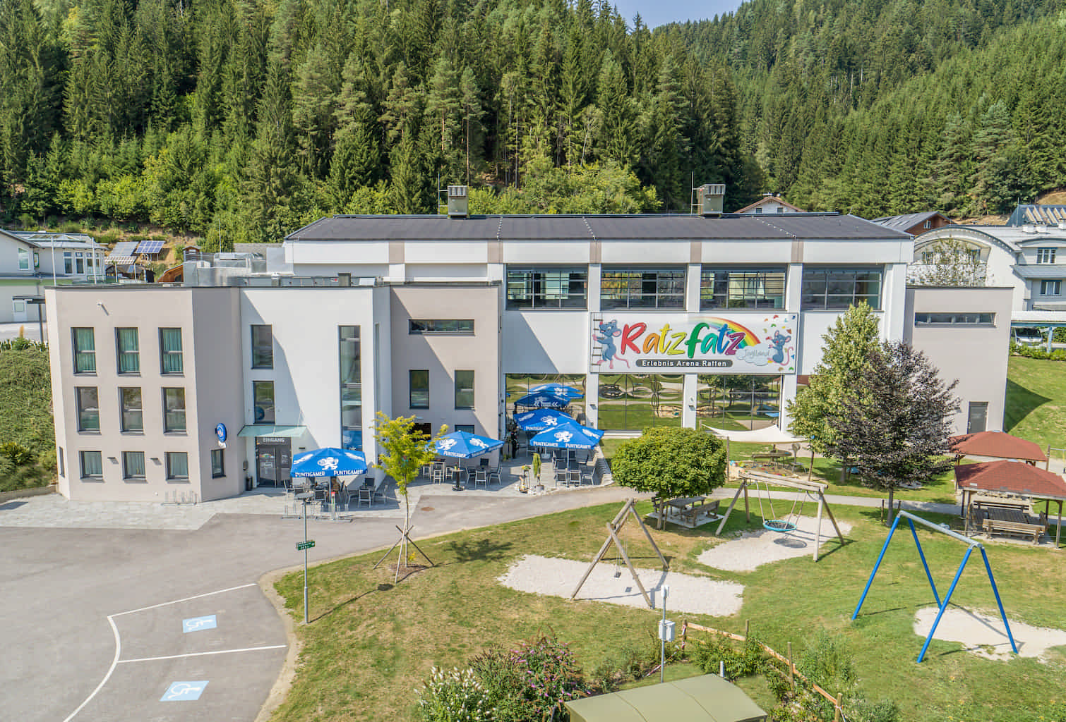 Außenaufnahme der Ratzfatz Erlebnisarena. Im Außenbereich des zweigeschoßigen Gebäudes finden sich ein Gastgarten, ein Spielplatz mit Schaukeln sowie ein Pavillon