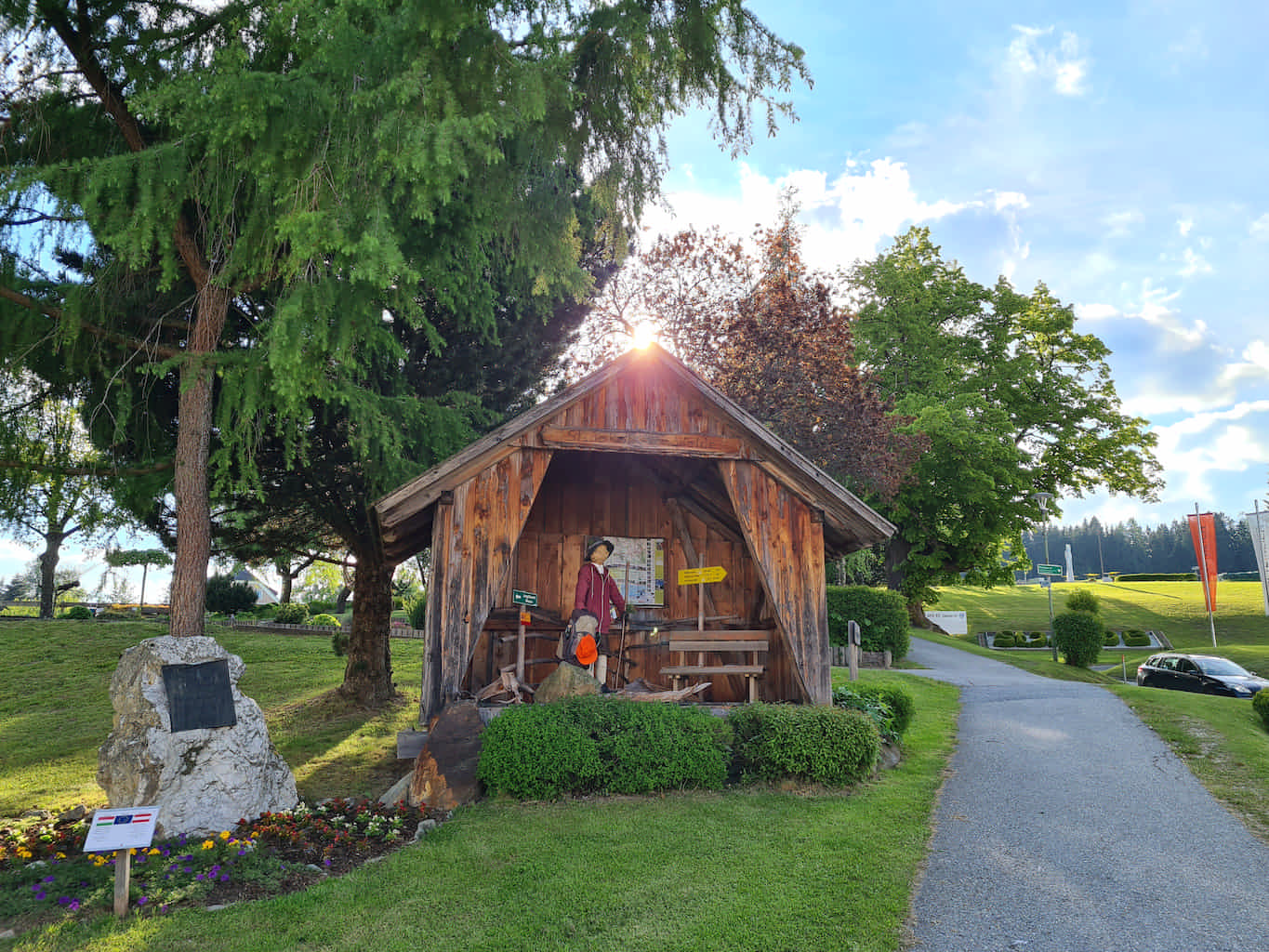 Kleine an einer Seite offene Holzhütte mit Skifahrerfigur und Wegweiser. Daneben befindet sich ein steinernes Denkmal unter einem Baum