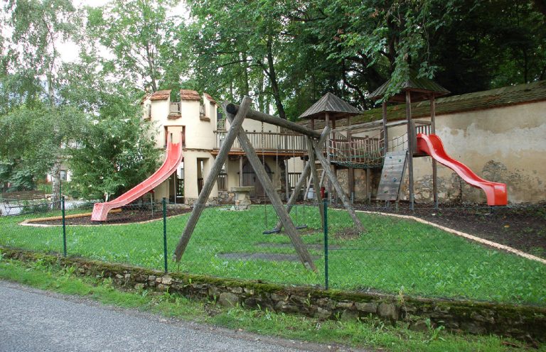 Das Bild zeigt einen Spielplatz in Burgoptik mit zwei Rutschen und zwei Schaukeln sowie weiteren Geräten